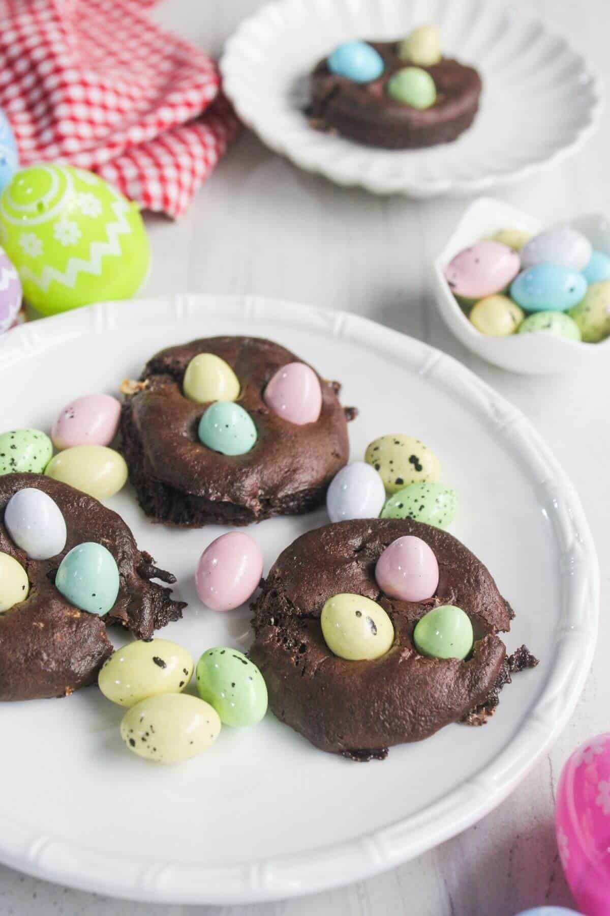 Chocolate cookies topped with pastel candy eggs are arranged on a white plate, with extra candy eggs and decorated Easter eggs nearby.