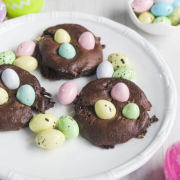 Three chocolate cake mix cookies topped with pastel candy eggs are arranged on a white plate, with additional candy eggs scattered nearby.