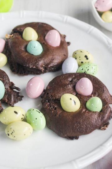 Three chocolate cake mix cookies topped with pastel candy eggs are arranged on a white plate, with additional candy eggs scattered nearby.