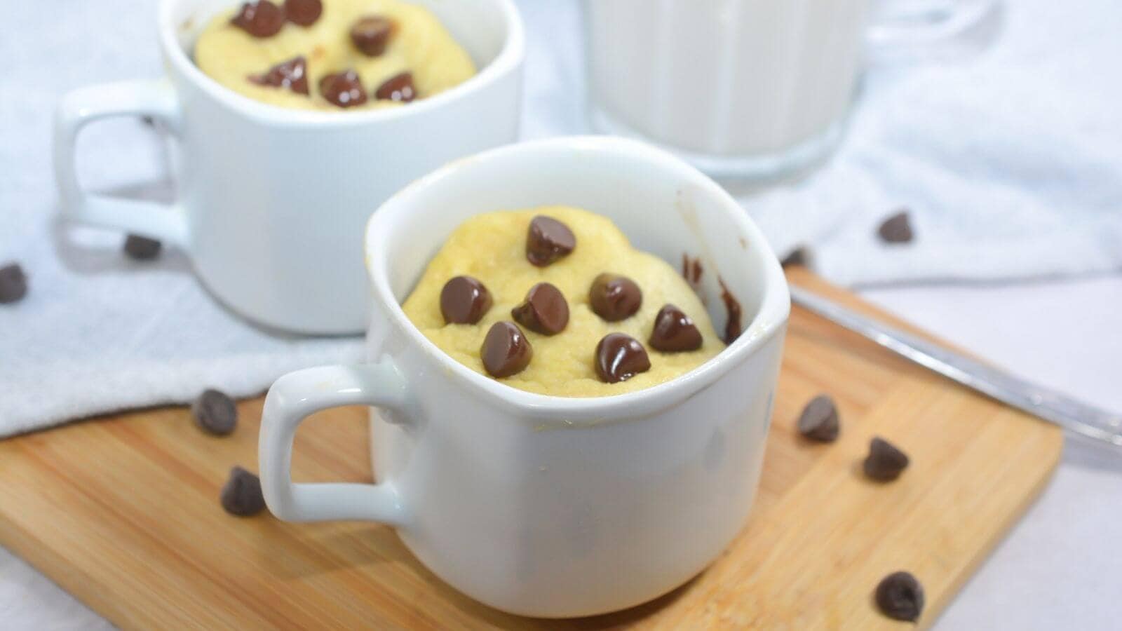 Two white mugs with chocolate chip mug cakes sit on a wooden board, with chocolate chips scattered around and a spoon nearby.
