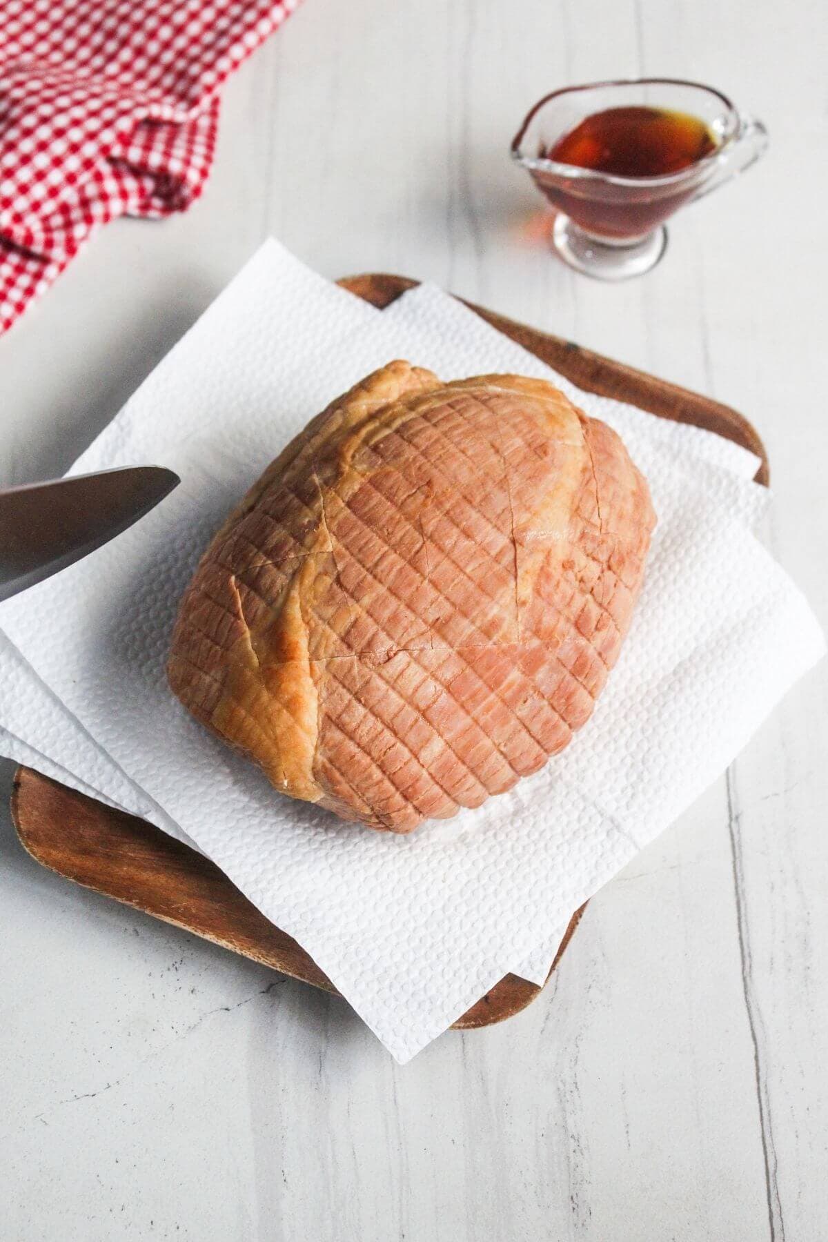 A cooked ham rests on paper towels atop a wooden tray, with a small glass pitcher of sauce and a red checkered cloth in the background.