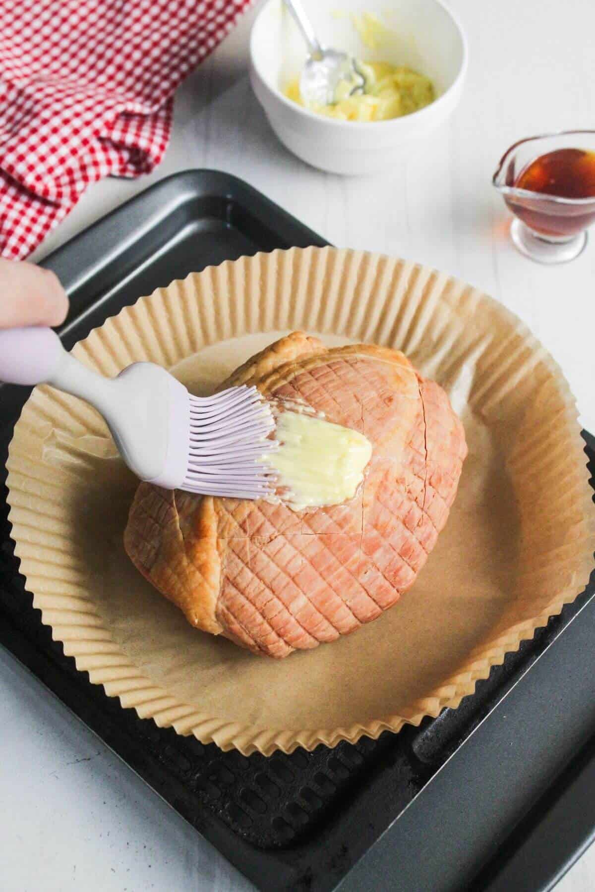 A hand brushes glaze over a scored, uncooked ham on parchment paper in a baking tray. A bowl of glaze and a small pitcher of liquid are nearby.