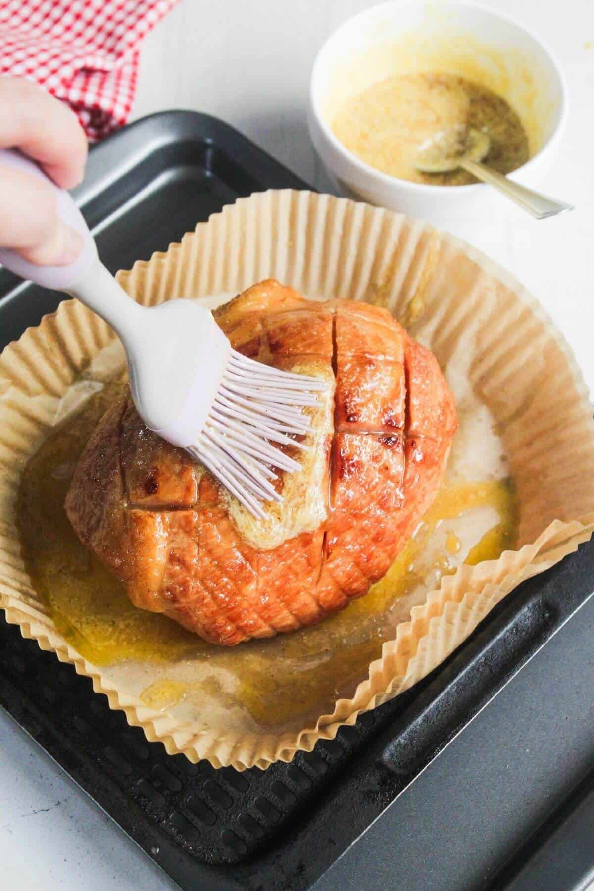 A hand uses a silicone brush to glaze a baked ham on parchment paper in a roasting pan, with a bowl of sauce nearby.