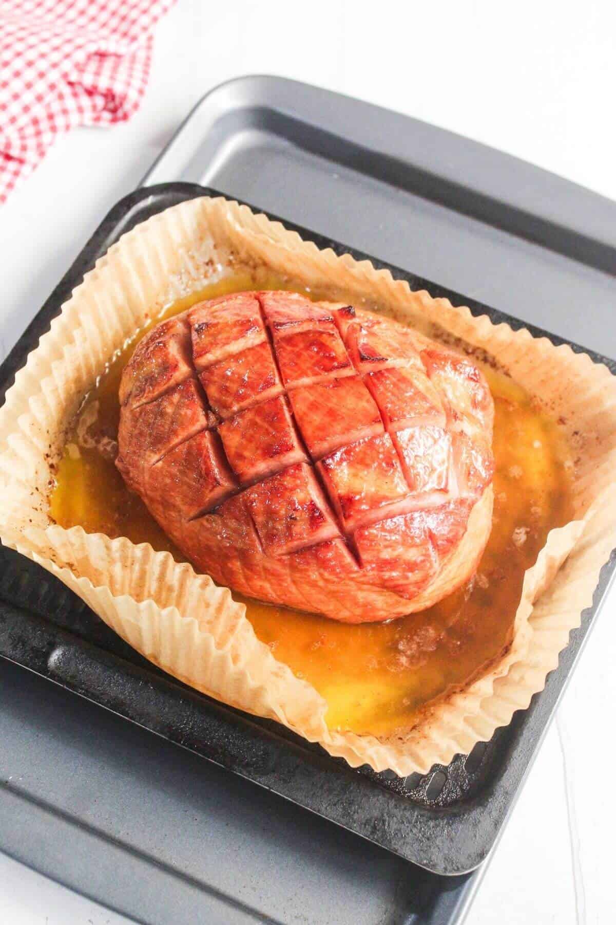 A cooked ham with a crosshatch pattern sits in a parchment-lined baking tray with juices pooled at the bottom.