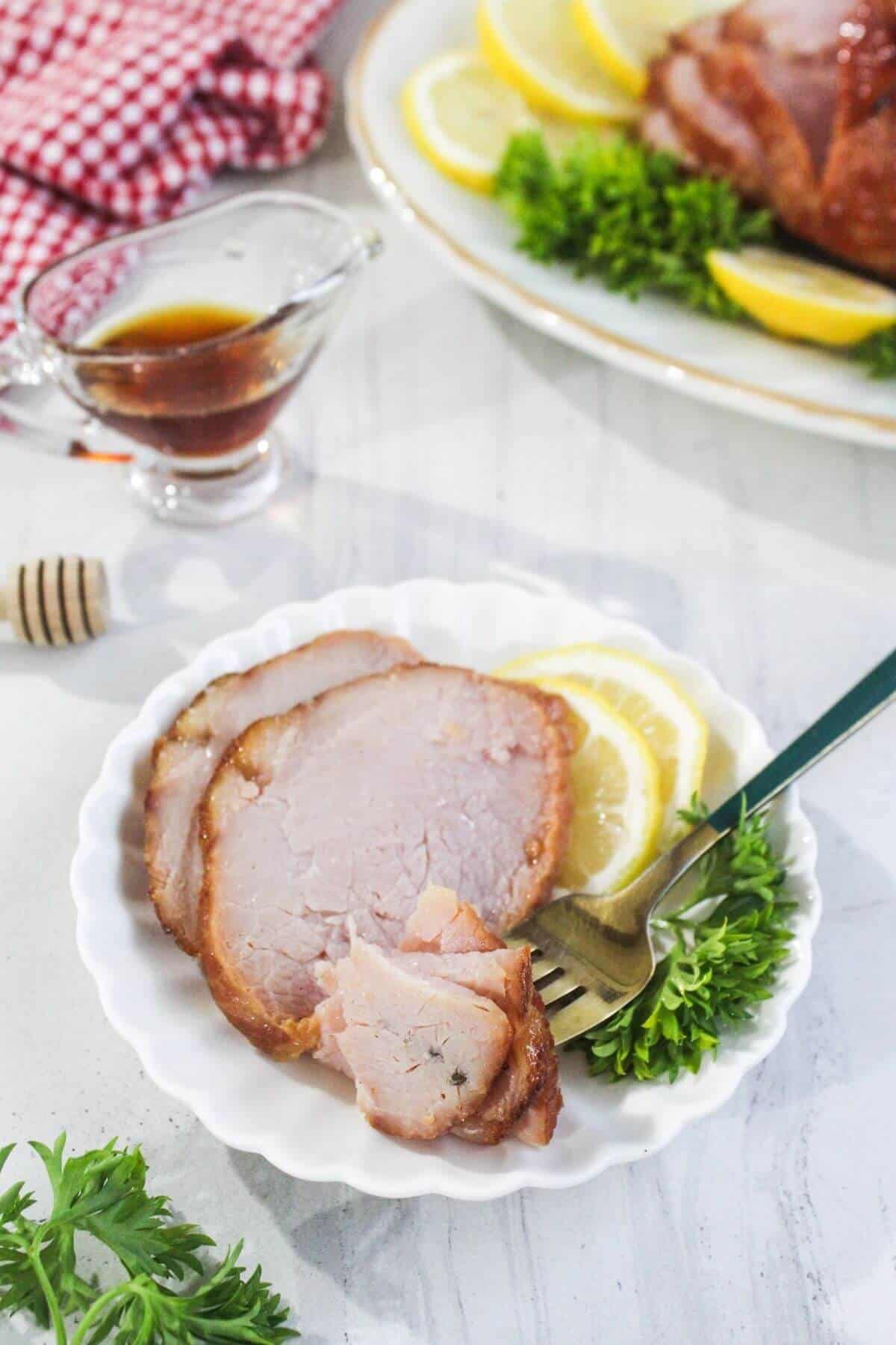 Three slices of cooked ham served on a white plate with lemon slices and parsley, with a fork, sauce, and a platter of ham in the background.