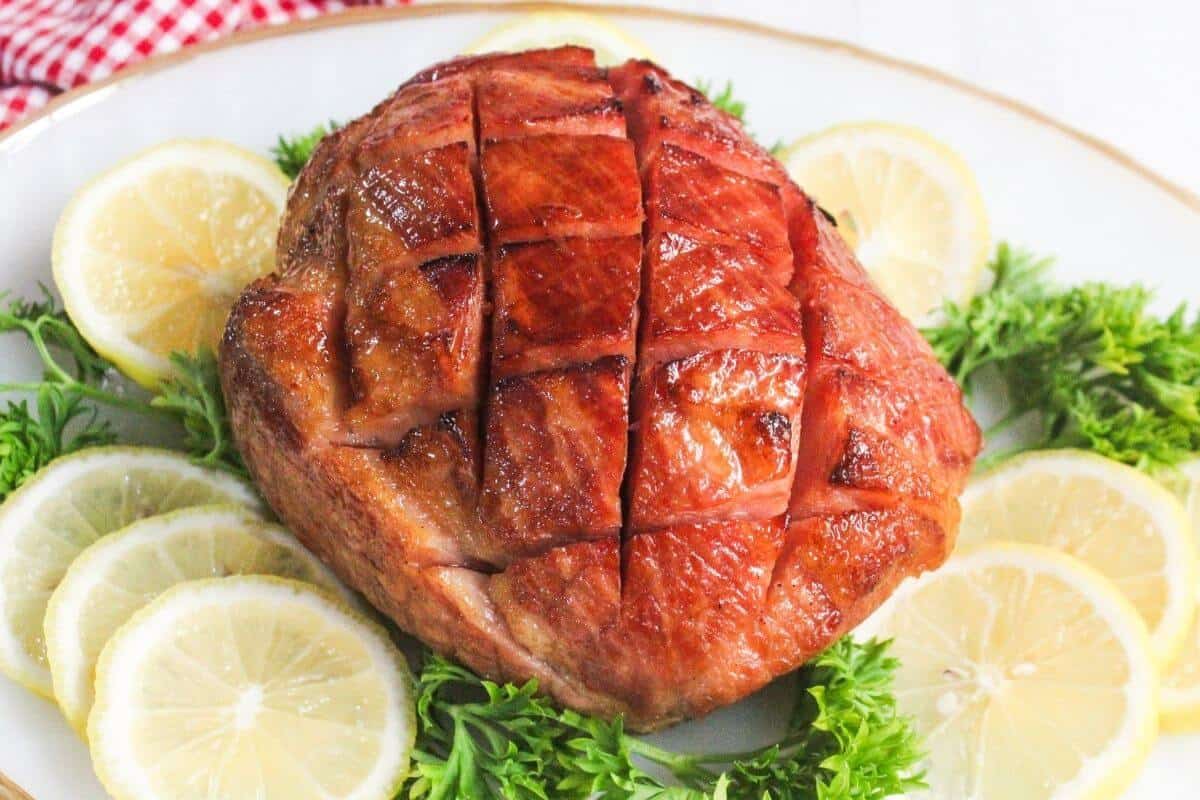 A cooked ham with a crosshatch pattern sits on a platter, surrounded by lemon slices and parsley.