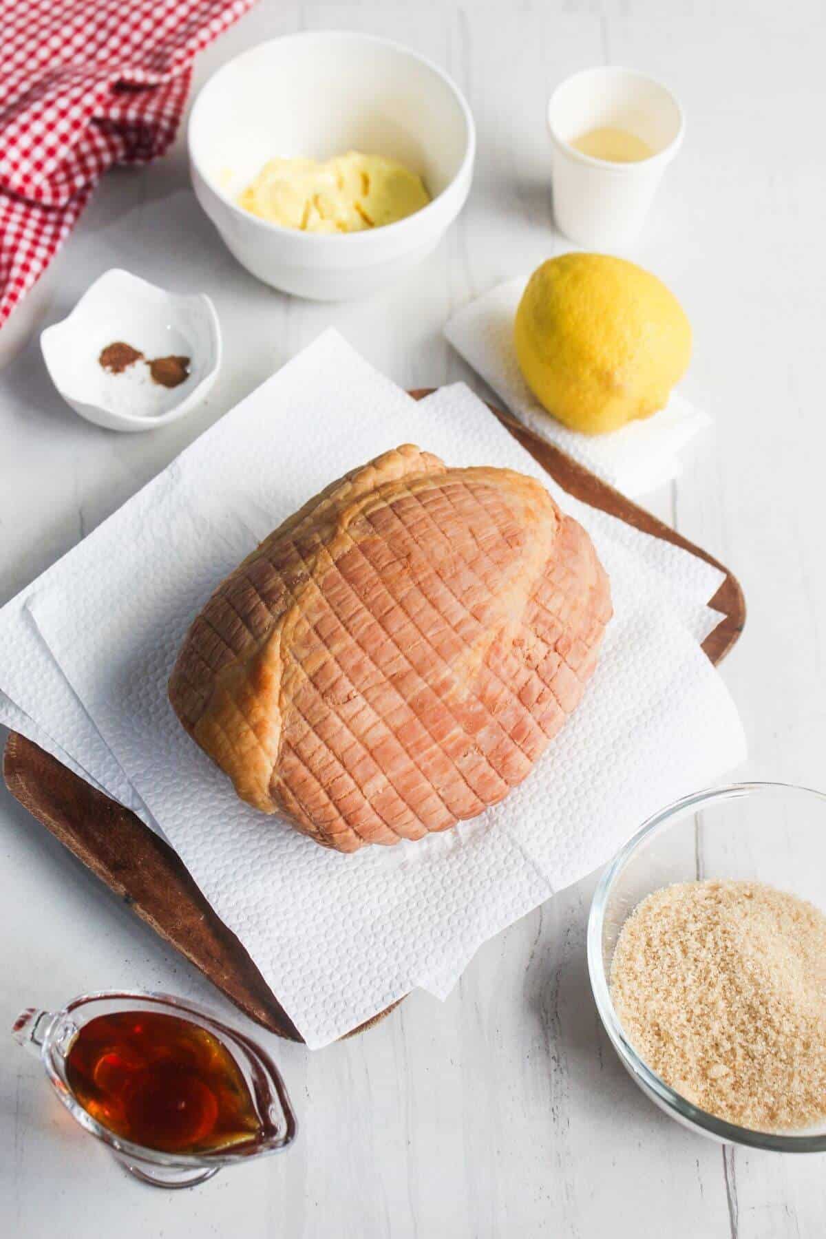 A cooked ham rests on paper towels on a wooden tray, surrounded by bowls of breadcrumbs, mustard, spices, a lemon, and a small pitcher of liquid on a white table.