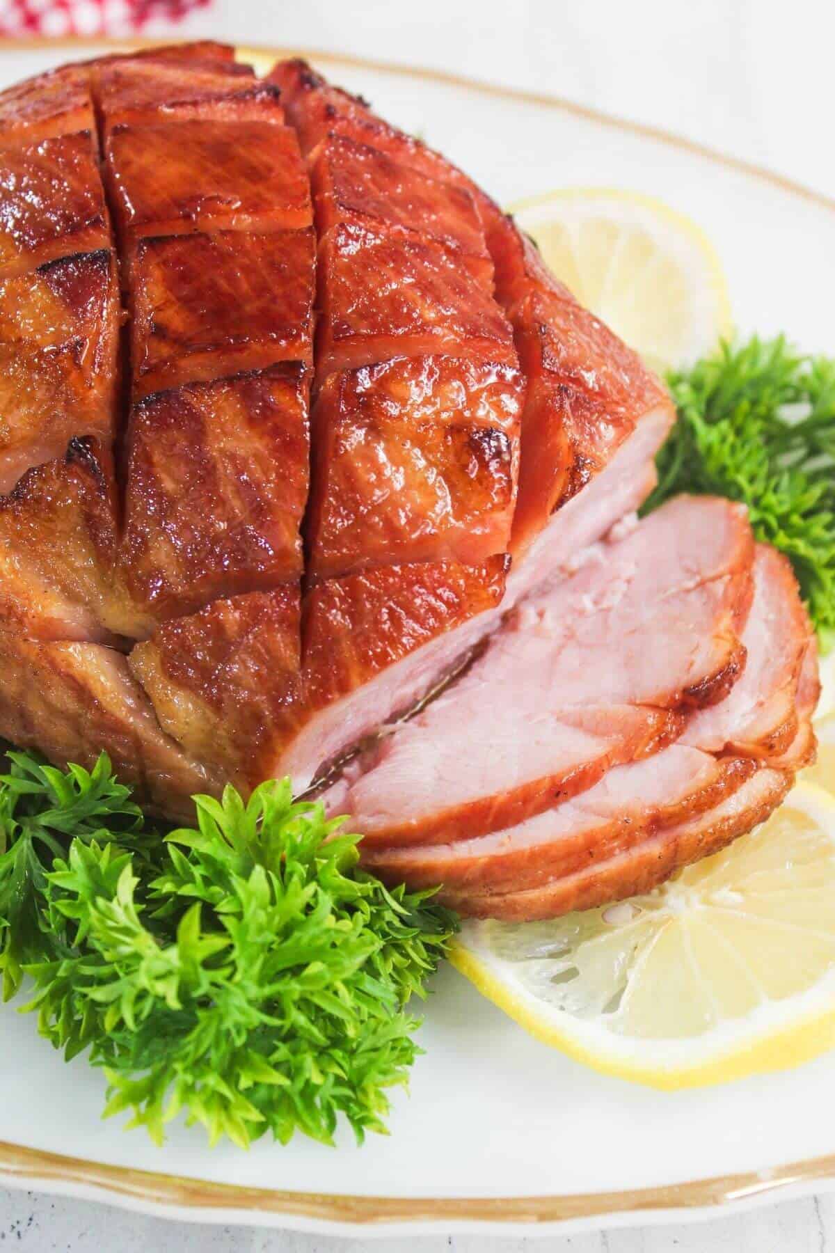 A glazed, sliced ham garnished with fresh parsley and lemon slices on a white plate.