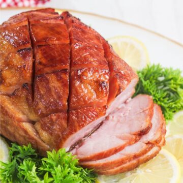 A sliced glazed ham with a crosshatch pattern, garnished with parsley and lemon slices on a white plate.