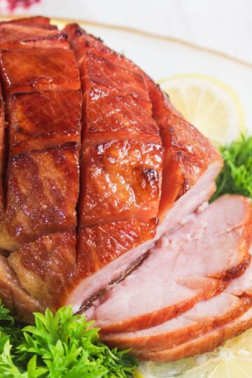A sliced glazed ham with a crosshatch pattern, garnished with parsley and lemon slices on a white plate.