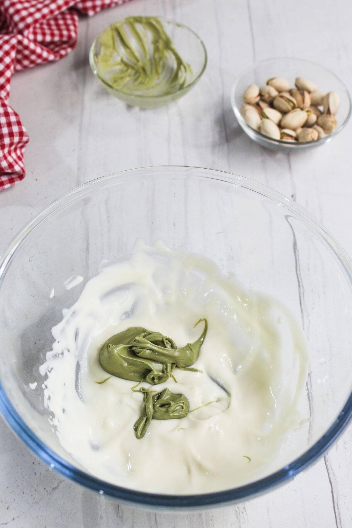A glass bowl with white yogurt or cream and green paste being mixed, a small bowl of pistachios, and a red checkered cloth on a white surface.