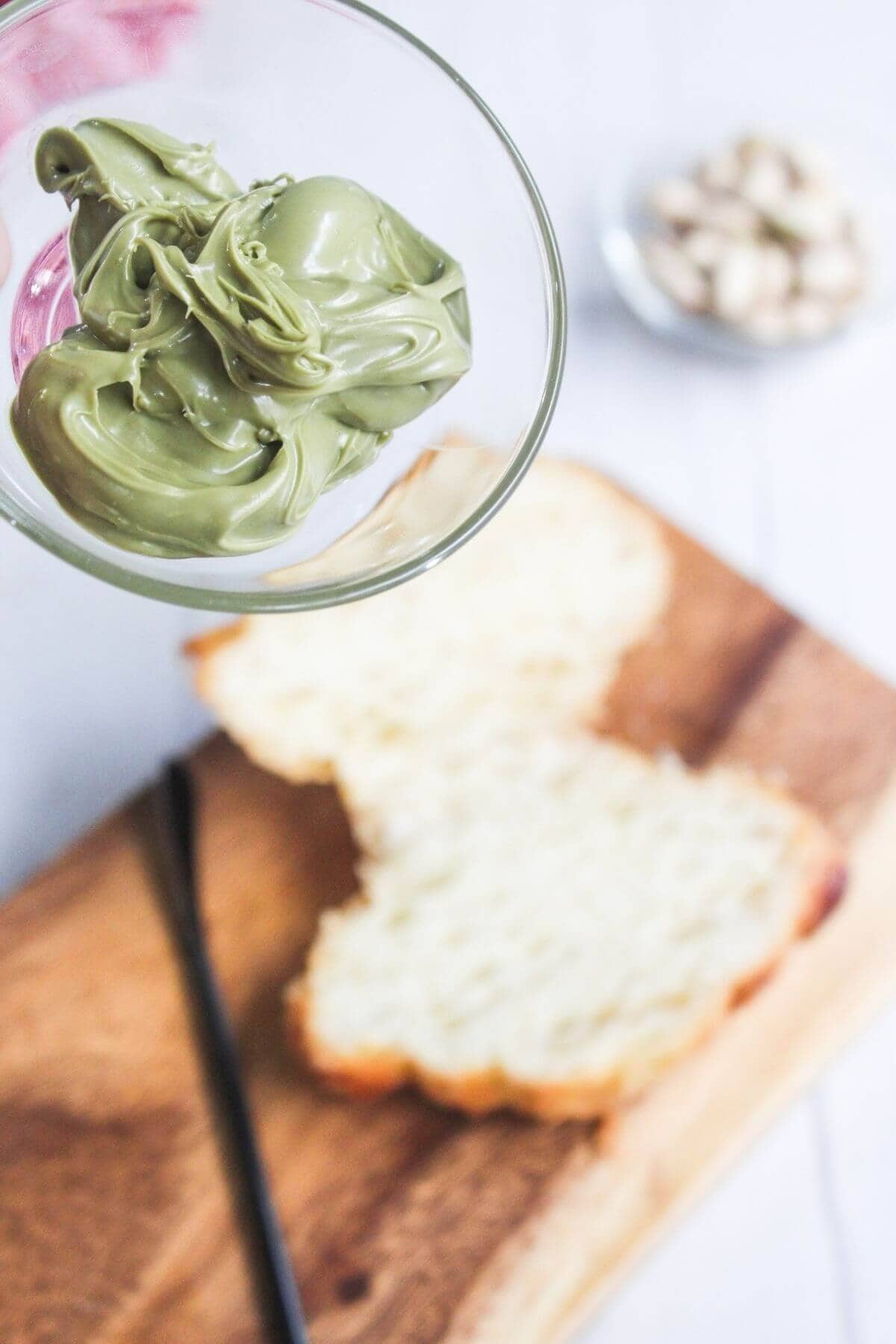 A bowl of green pistachio spread held above a slice of bread on a wooden board, with a butter knife and a small bowl of pistachios in the background.