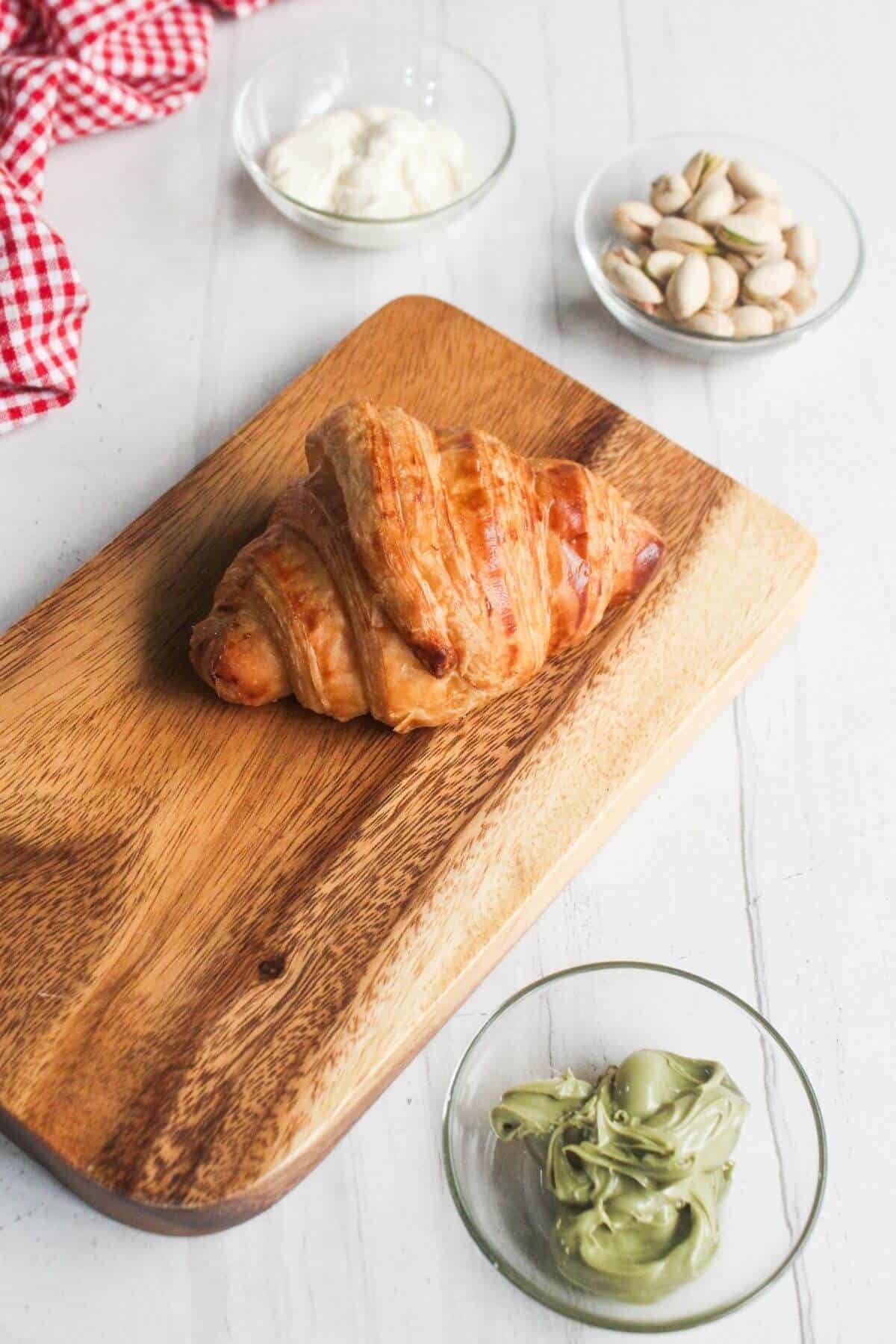 A croissant on a wooden board, with small glass bowls containing yogurt, pistachios, and green pistachio cream nearby on a white surface.