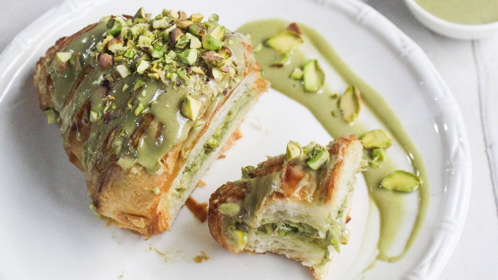 A croissant topped and filled with green pistachio cream and chopped pistachios, served on a white plate with extra pistachio cream and nuts on the side.