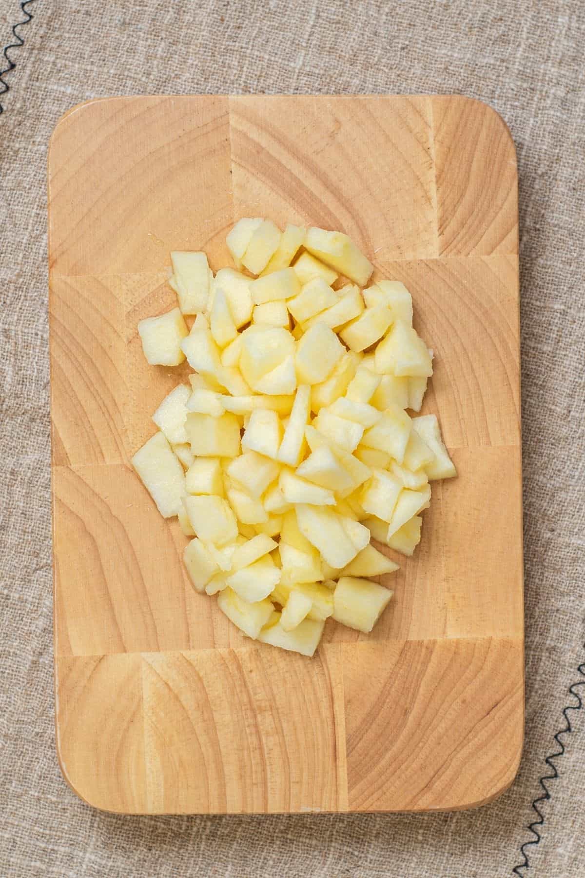 Diced apple pieces arranged in the center of a wooden cutting board on a textured fabric surface.