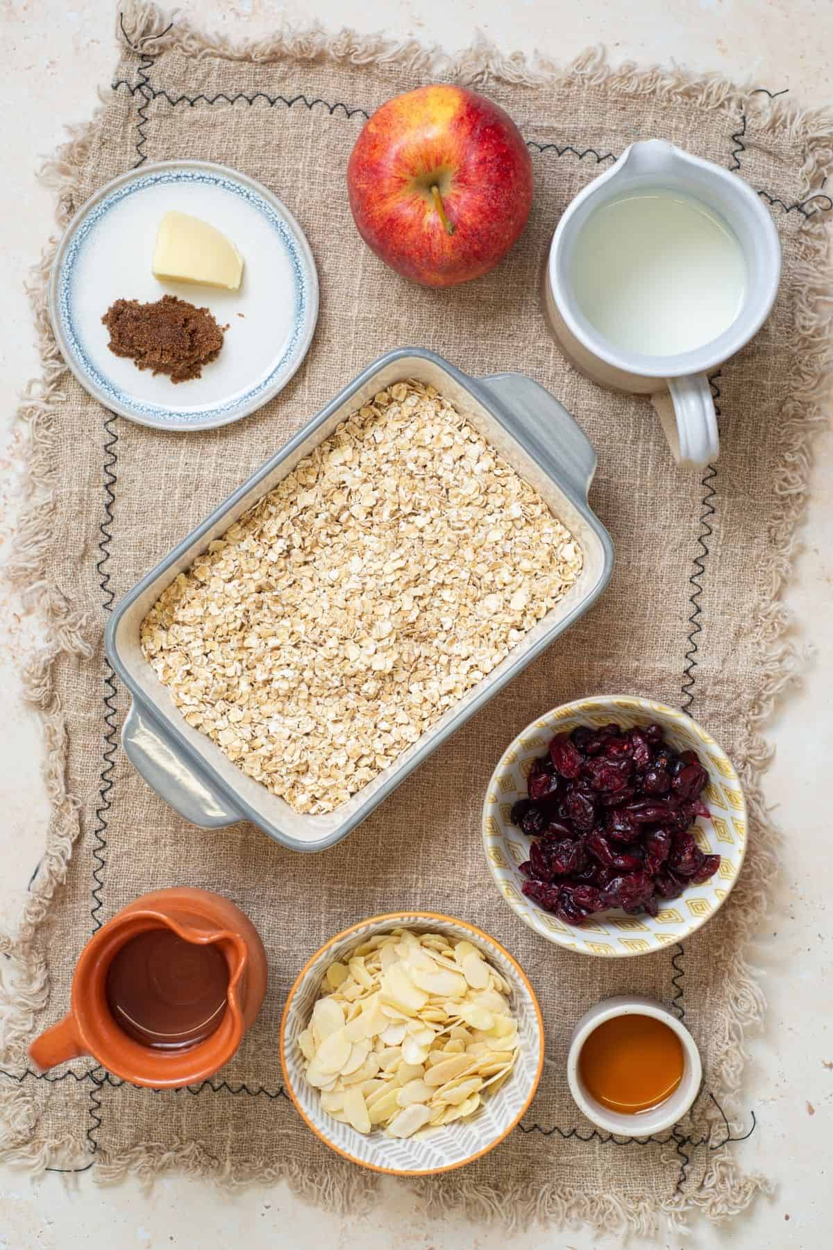 A flatlay of ingredients for baked oatmeal, including oats, milk, an apple, butter, brown sugar, dried cranberries, sliced almonds, syrup, and vanilla extract.