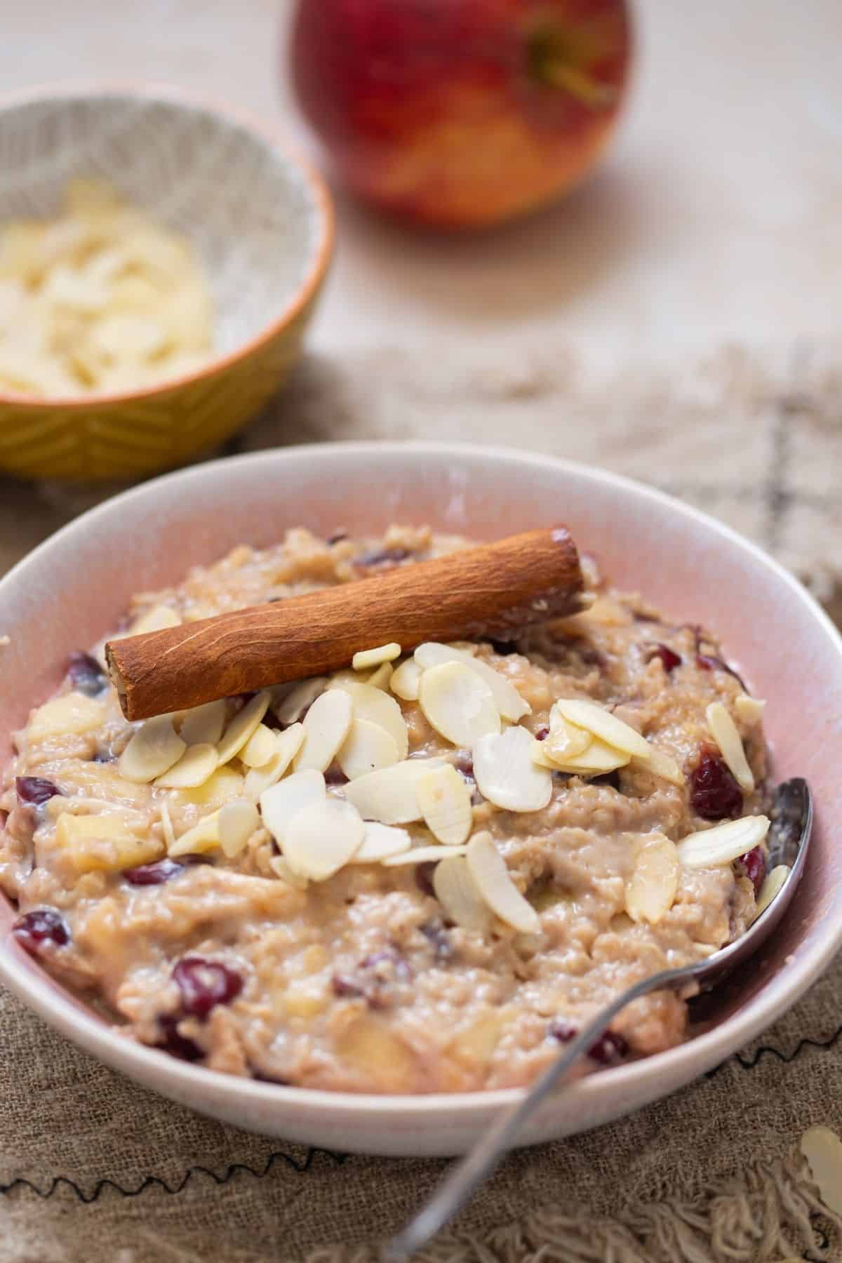 A bowl of oatmeal topped with sliced almonds and a cinnamon stick, with a spoon in the bowl and an apple and almond slices in the background.