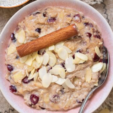 A bowl of oatmeal topped with sliced almonds, dried cranberries, and a cinnamon stick, with a spoon resting inside the bowl.