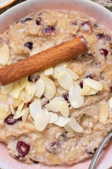 A bowl of oatmeal topped with sliced almonds, dried cranberries, and a cinnamon stick, with a spoon resting inside the bowl.