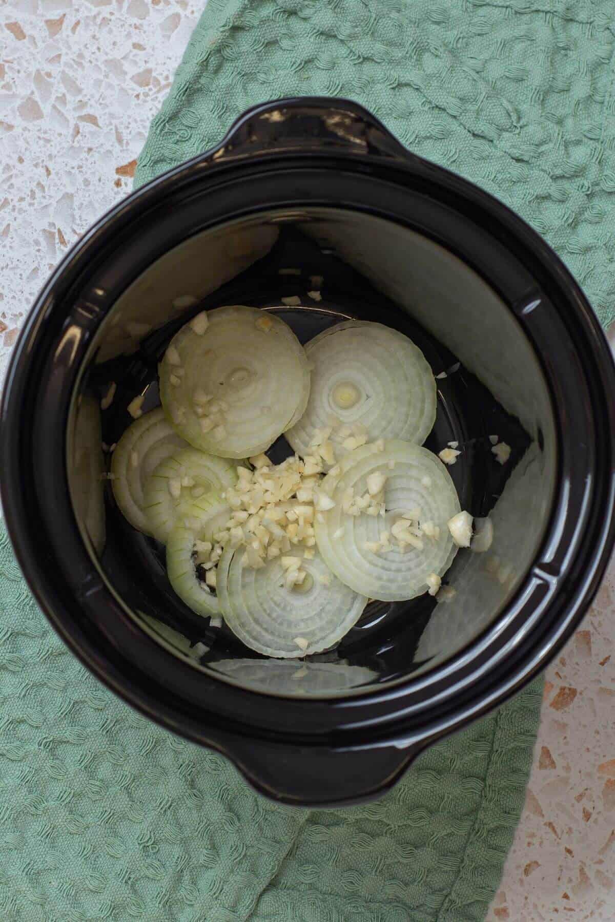 Sliced onions and minced garlic placed at the bottom of a black slow cooker, set on a green textured cloth.