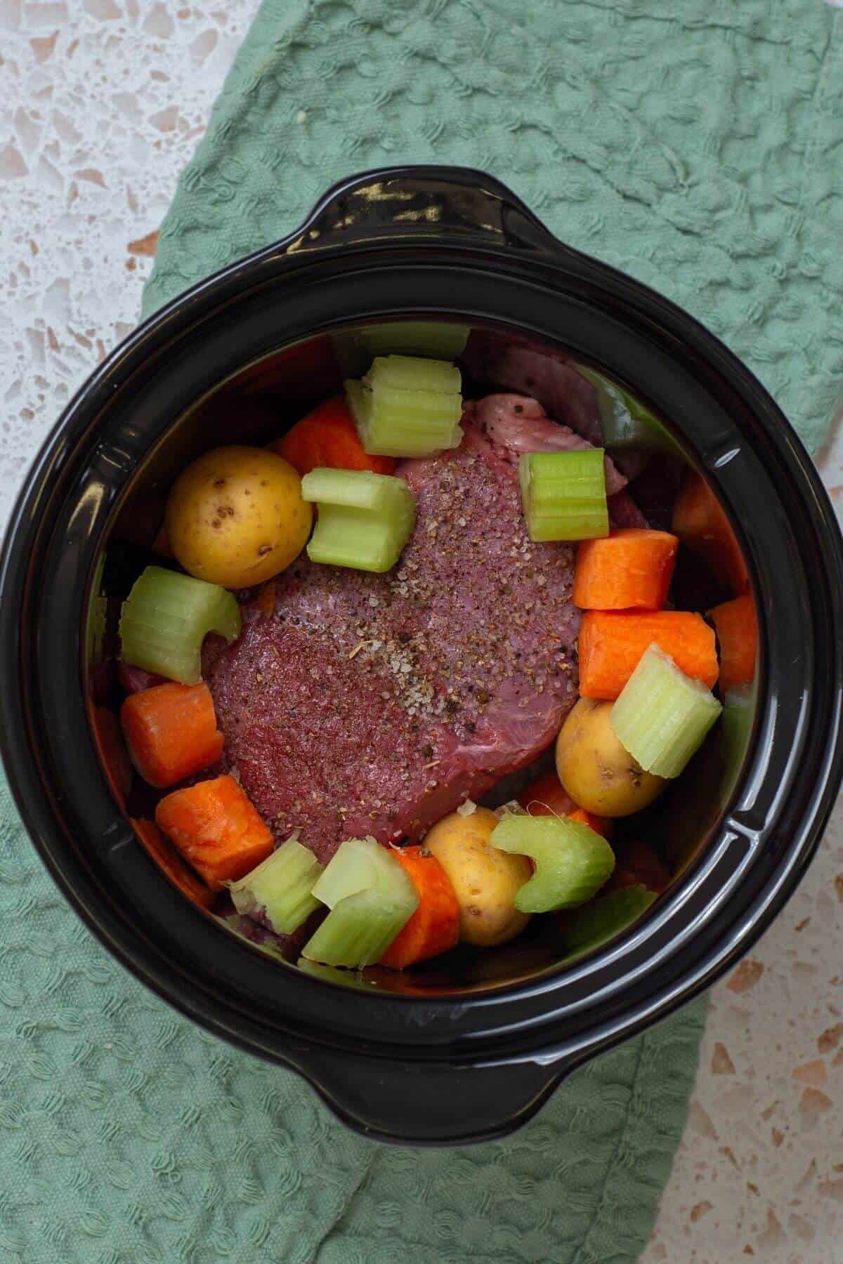 A raw beef roast with potatoes, carrots, and celery in a slow cooker, ready to be cooked, sits on a green textured cloth.