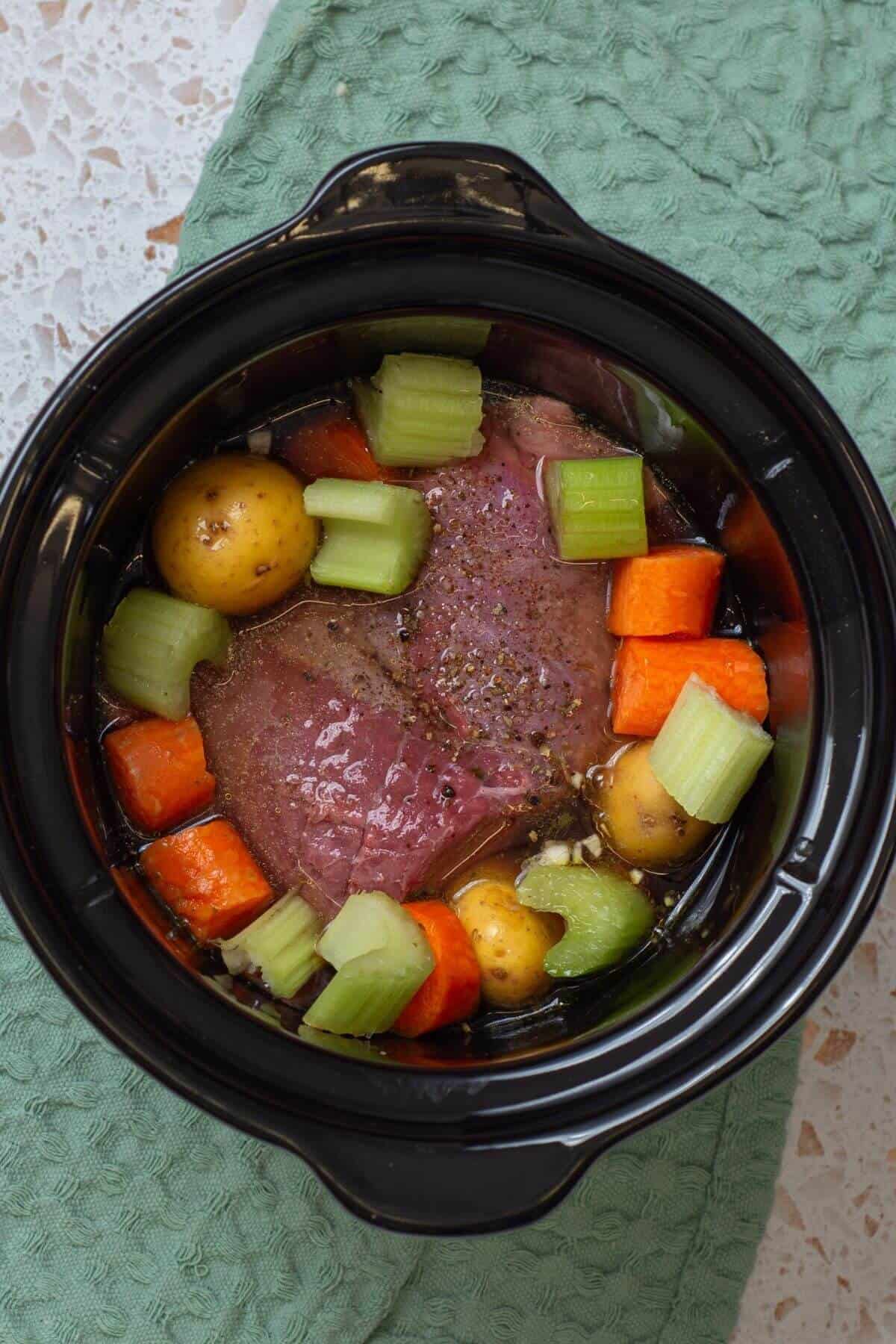 A slow cooker filled with raw beef, potatoes, carrots, and celery on a green cloth.