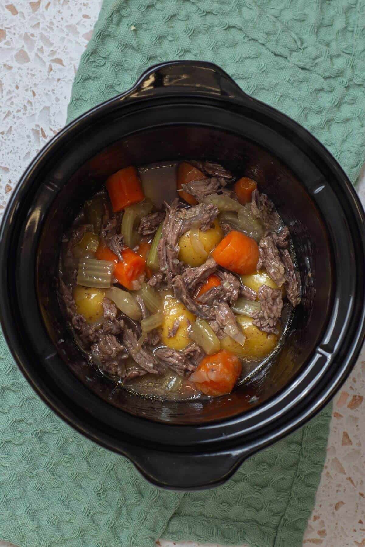 Overhead view of a beef stew with potatoes, carrots, and onions in a black slow cooker on a green textured cloth.