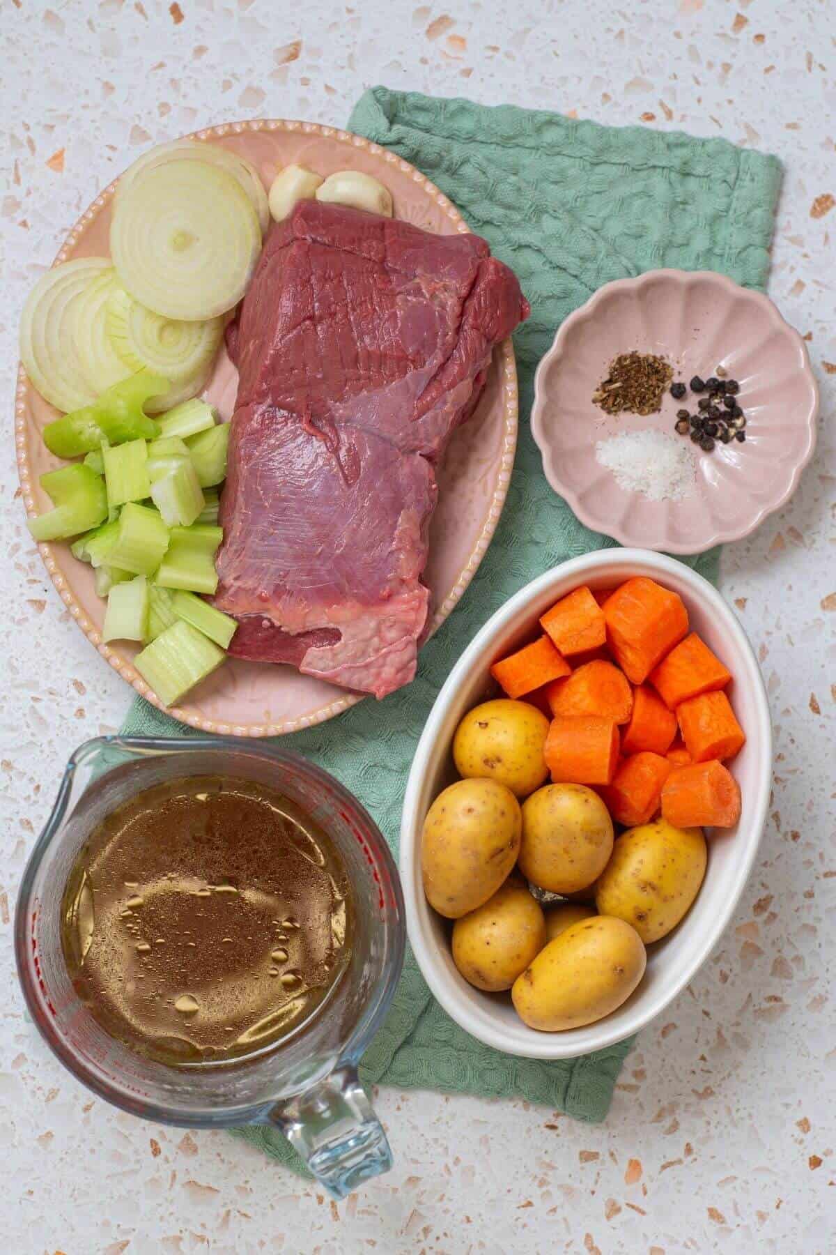 Raw ingredients including sliced onion, garlic, celery, a slab of beef, baby potatoes, chopped carrots, seasonings, and a measuring cup of broth arranged on a counter.