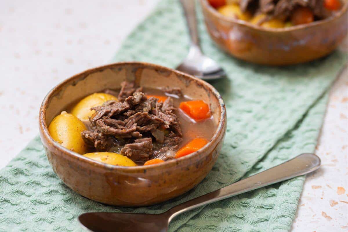 A bowl of beef stew with chunks of beef, potatoes, and carrots sits on a green textured cloth, with a spoon beside it and another bowl in the background.