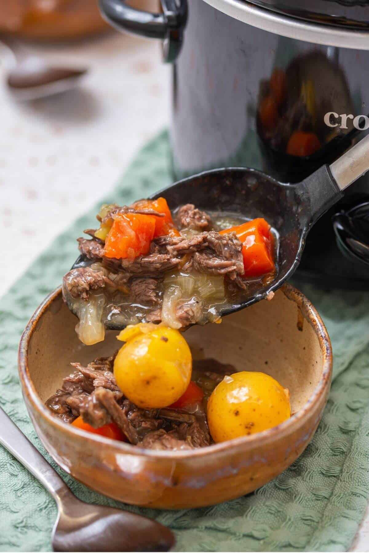 A ladle serving beef stew with potatoes, carrots, and onions from a slow cooker into a ceramic bowl.