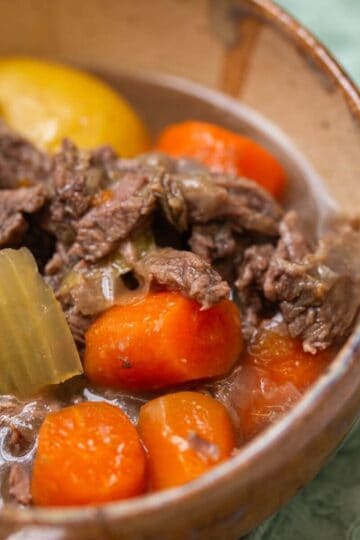 A bowl of beef stew with chunks of beef, carrots, potatoes, and celery in a brown ceramic bowl.