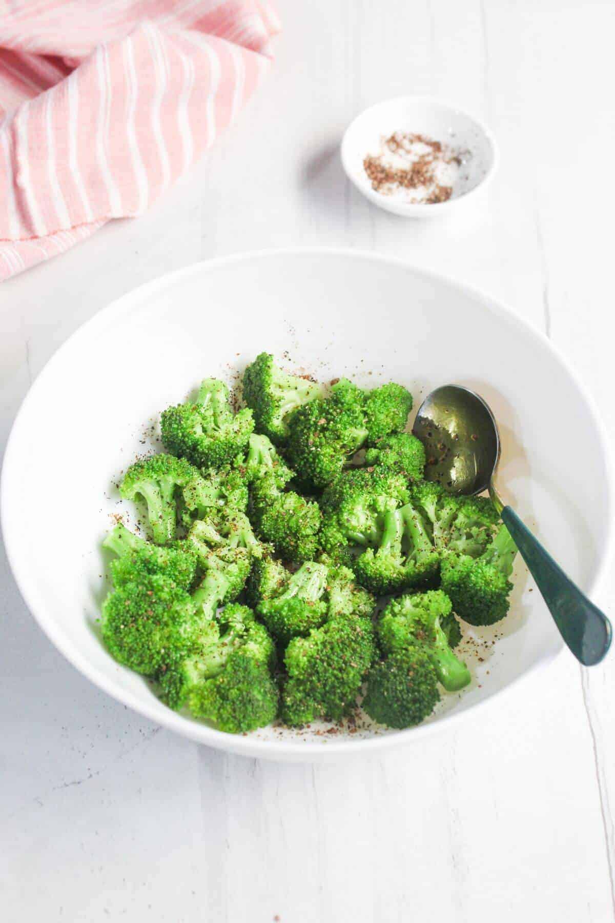 A white bowl filled with steamed broccoli florets topped with black pepper, with a spoon inside. A small dish of seasoning and a striped towel are in the background.