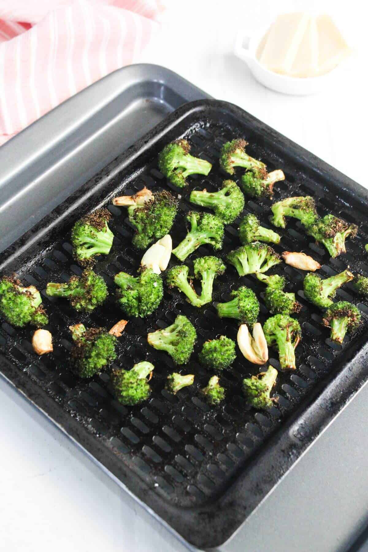Broccoli florets and garlic cloves roasted on a black grill pan, with a pink striped cloth and small bowl in the background.