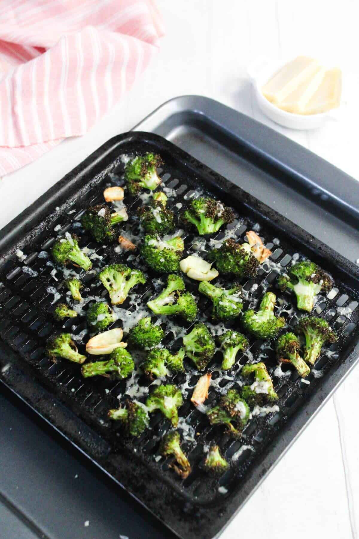 Roasted broccoli florets and garlic cloves on a black grill pan, sprinkled with grated cheese, with a striped towel and a small bowl in the background.
