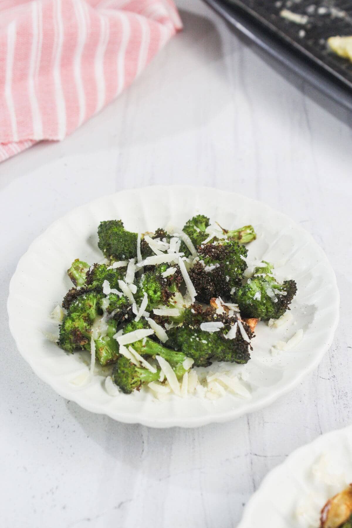A white plate with roasted broccoli florets topped with shredded cheese, set on a white surface with a pink striped cloth in the background.