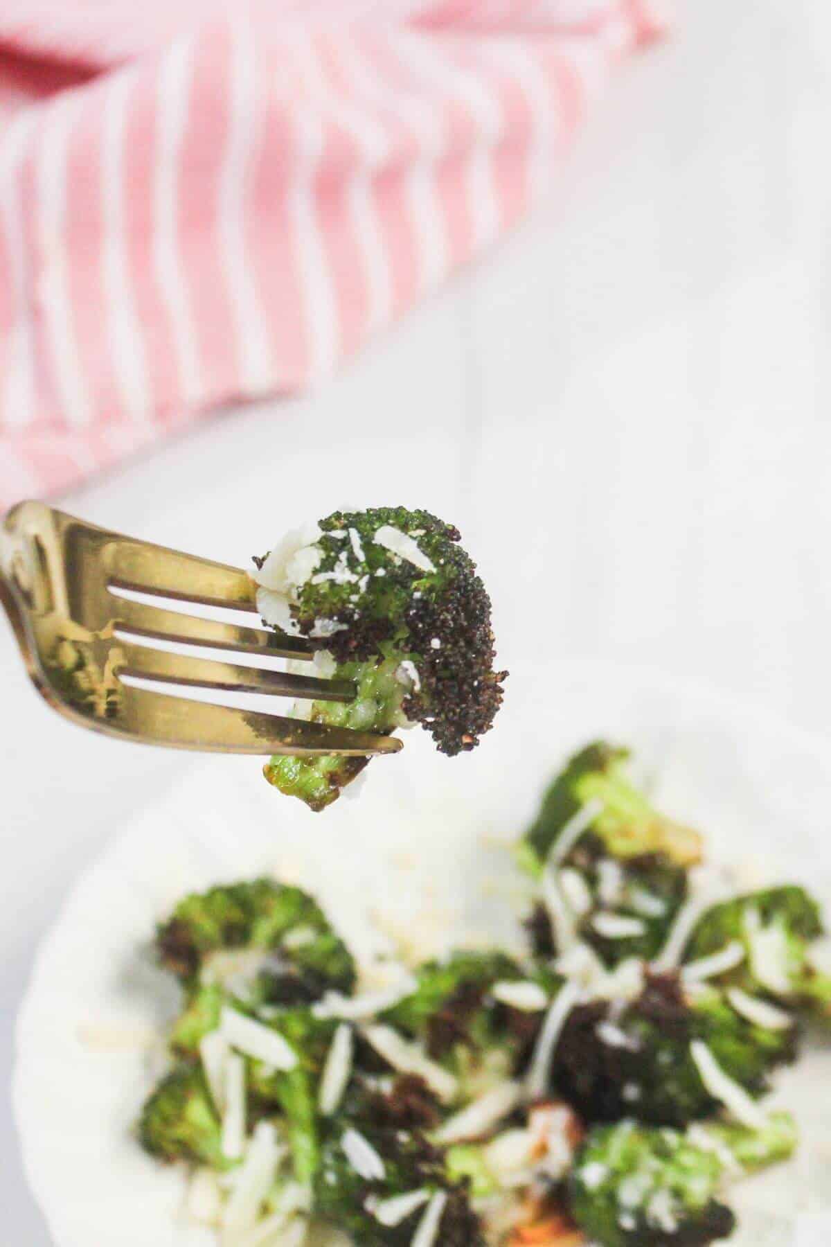 A close-up of a fork holding a piece of roasted broccoli with grated cheese, with more broccoli and cheese on a plate below. A pink striped cloth is in the background.
