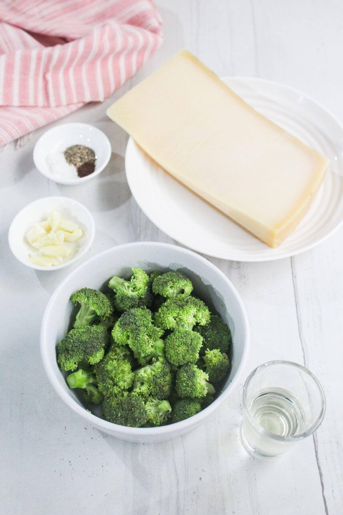 A bowl of broccoli florets, a glass of clear liquid, a plate with a block of cheese, a small dish of sliced garlic, and a dish of salt and pepper on a white surface.