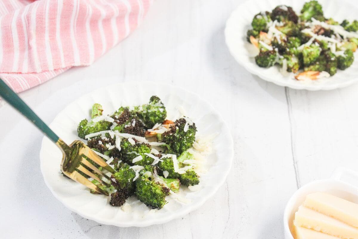 A white plate with roasted broccoli topped with shredded cheese, a fork, and another similar plate and a bowl with cheese in the background.