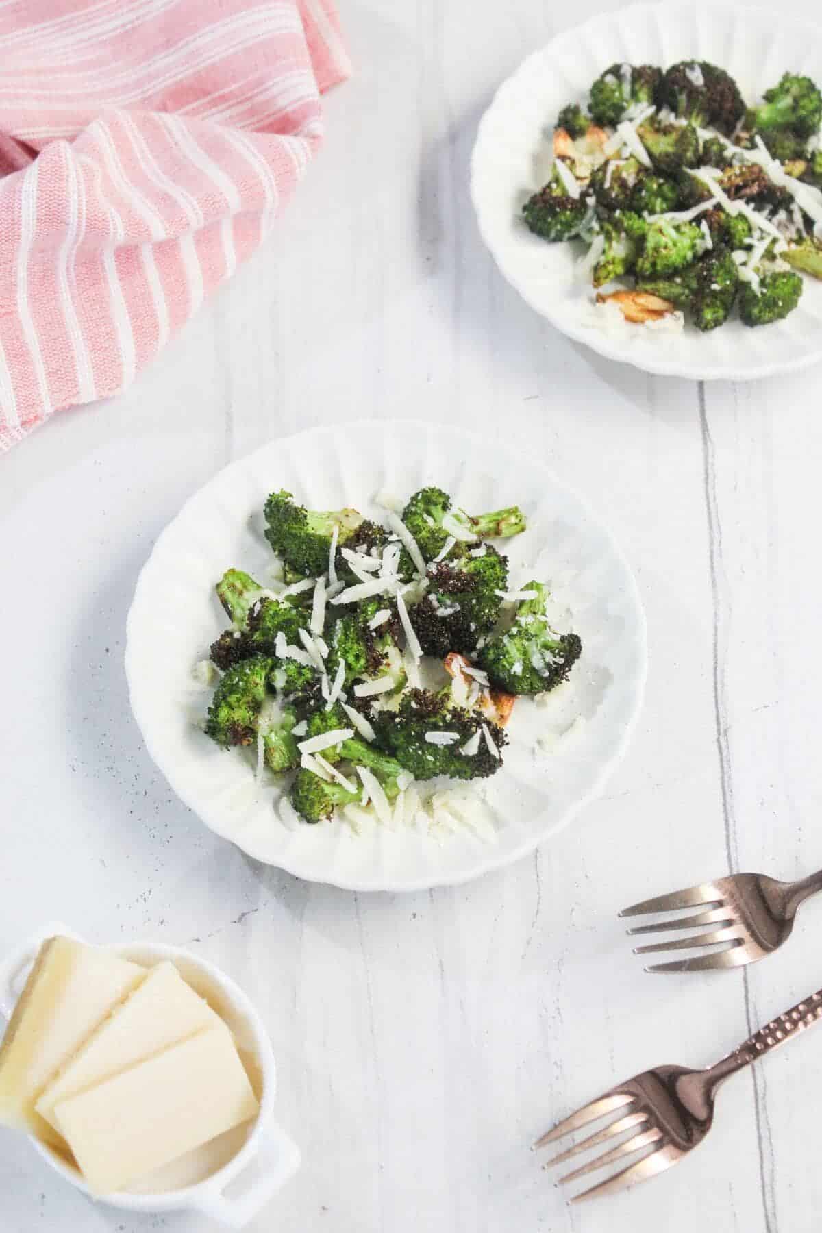 Two white plates with roasted broccoli topped with shredded cheese on a white surface, next to a pink striped cloth, forks, and a small dish of cheese slices.