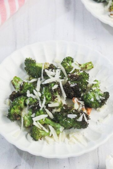 A white plate with roasted broccoli florets topped with shredded cheese, set on a white surface with a pink striped cloth partly visible.