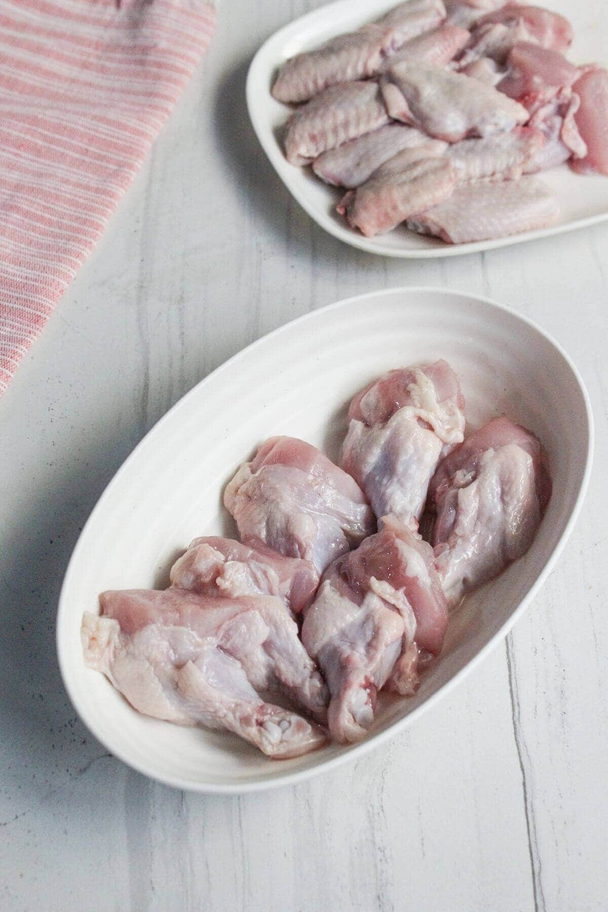 Two white dishes with raw chicken wings on a light-colored surface, one dish in the foreground and one in the background near a pink-striped towel.