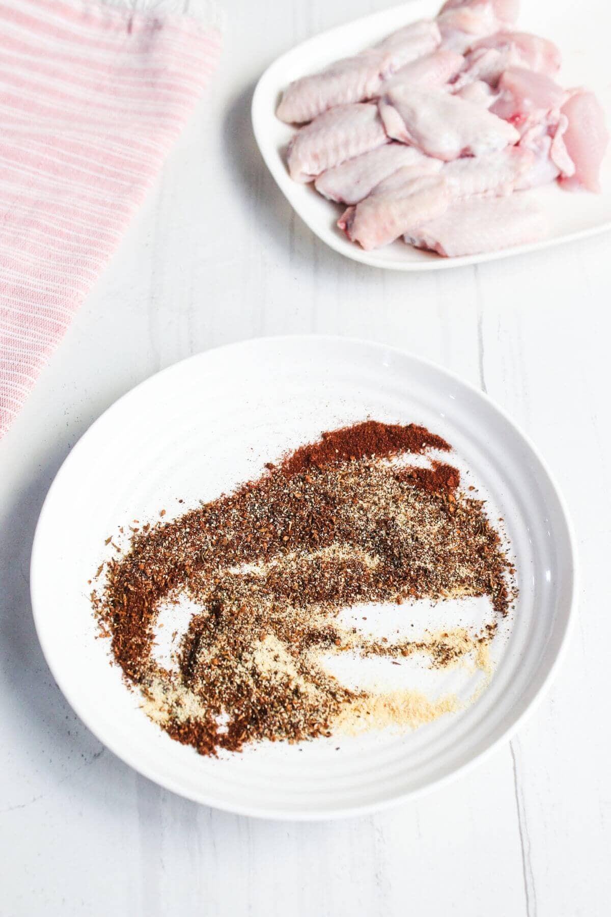 A white plate with assorted dry spices, with a plate of raw chicken wings and a pink-striped towel in the background.