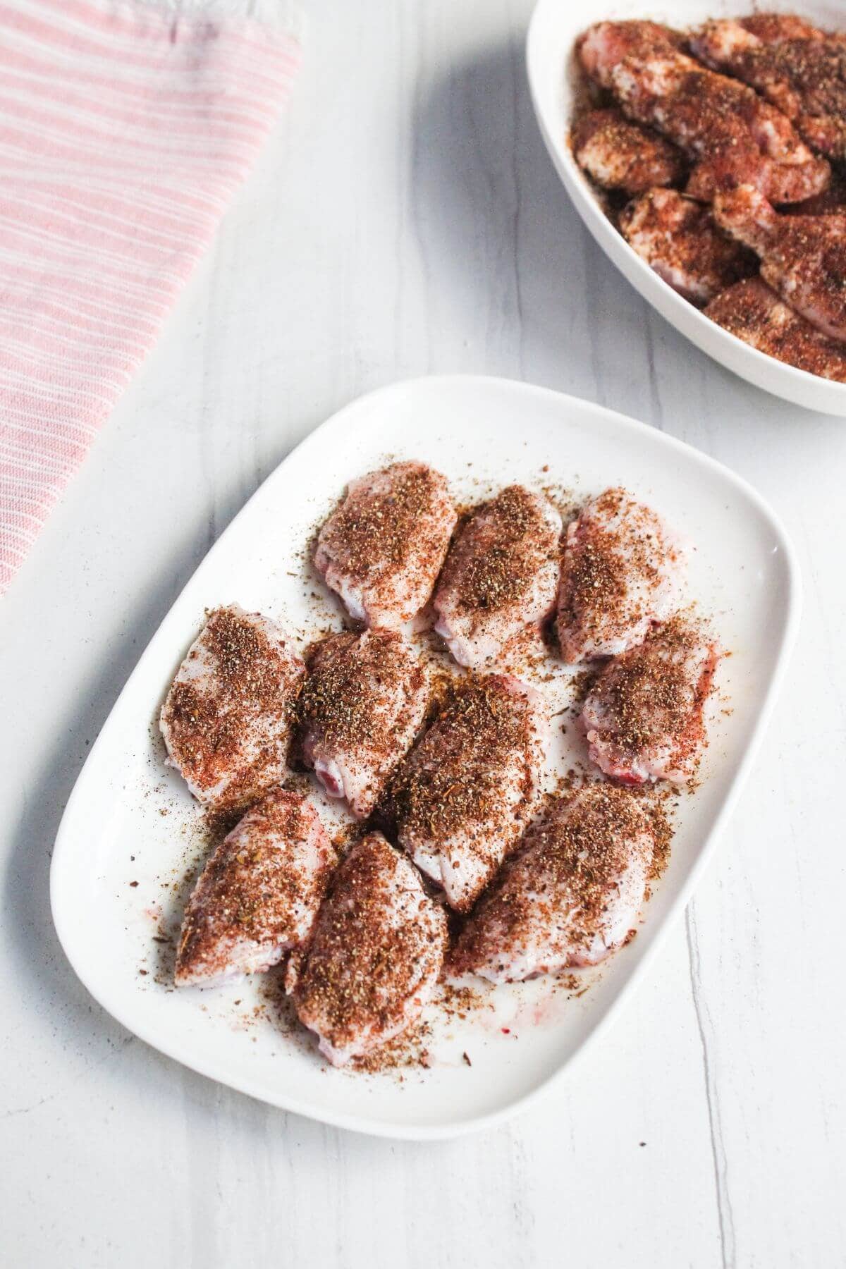 A white plate with raw chicken wings coated in seasoning, placed on a white surface; a bowl of more seasoned wings is in the background.