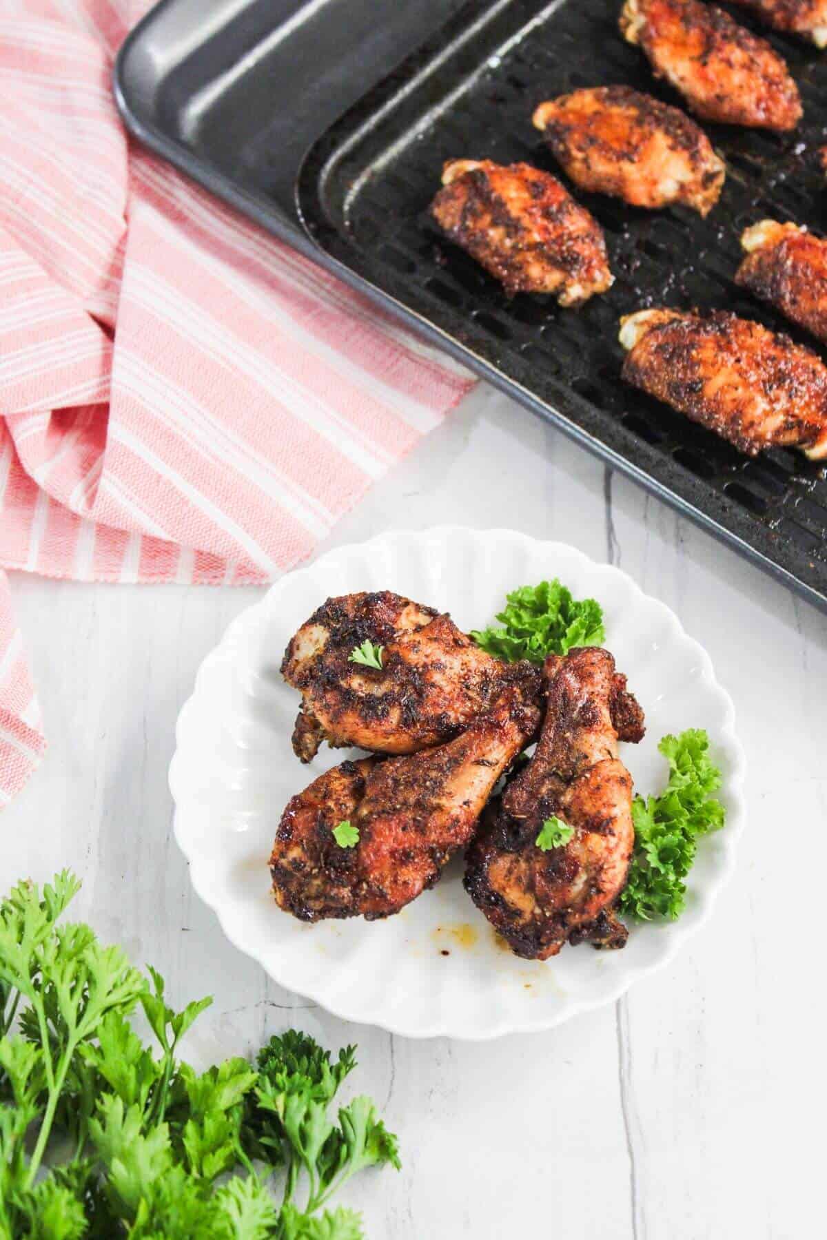 Three baked chicken drumsticks on a white plate with parsley garnish, next to a tray of additional chicken pieces, on a white surface with a pink striped cloth.