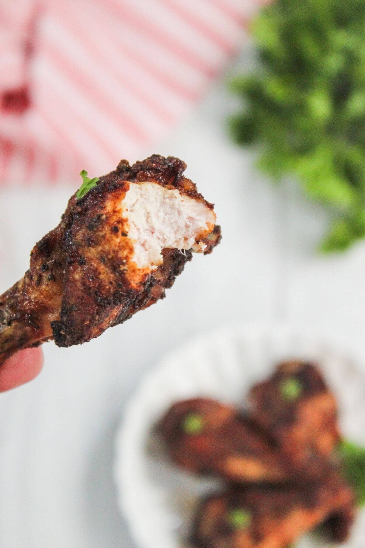 A close-up of a cooked chicken drumstick with a bite taken out, held in hand, with more drumsticks on a plate in the background.