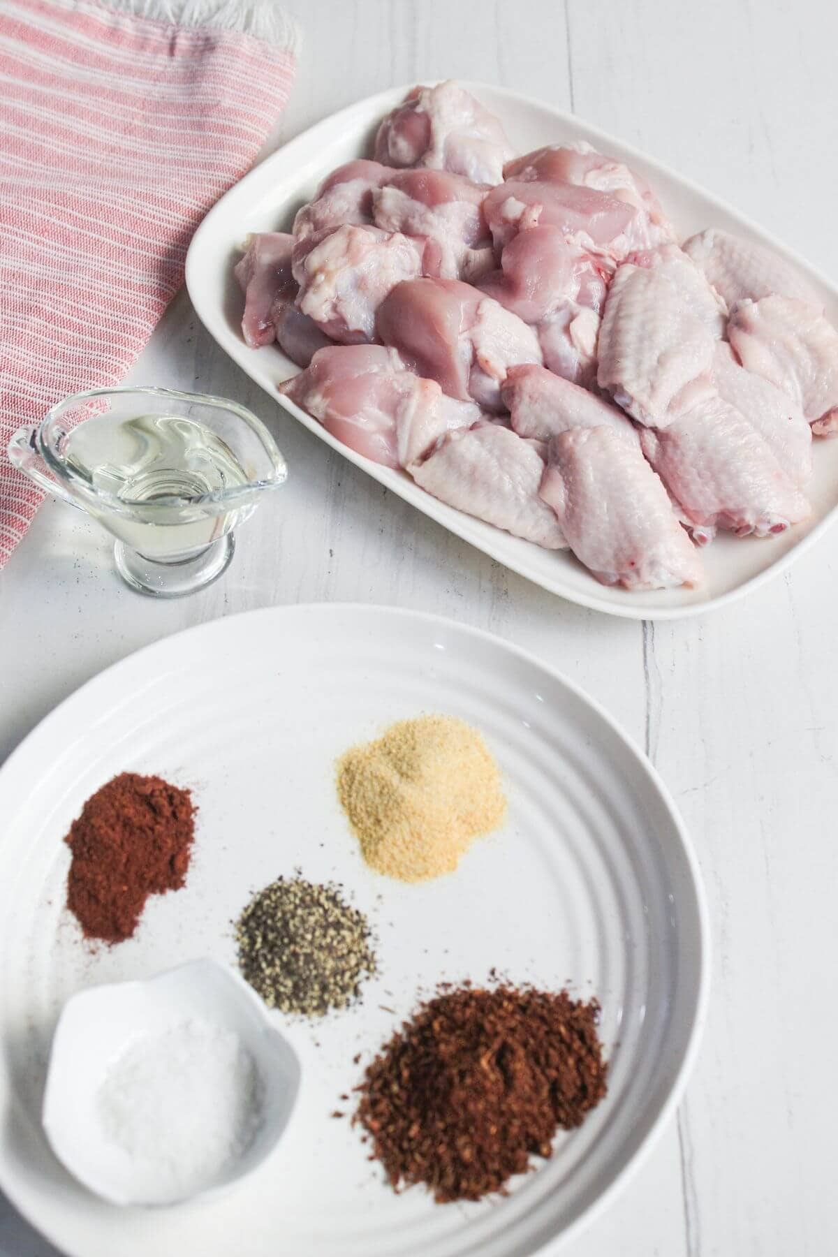 Raw chicken wings on a white platter, a small glass container of oil, and a plate with spices including salt, black pepper, paprika, chili powder, and garlic powder.