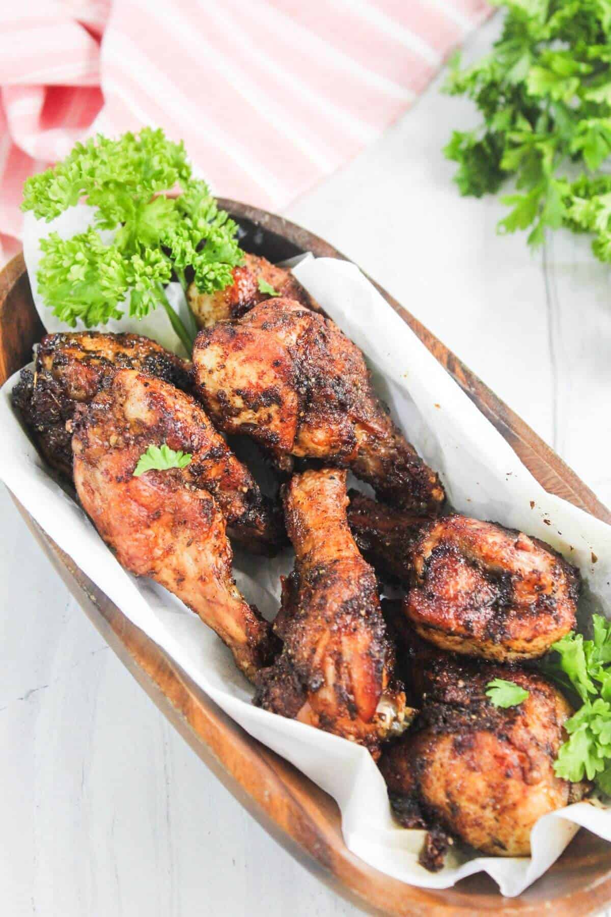 Grilled chicken drumsticks with a dark seasoning served on parchment in a wooden dish, garnished with fresh parsley.