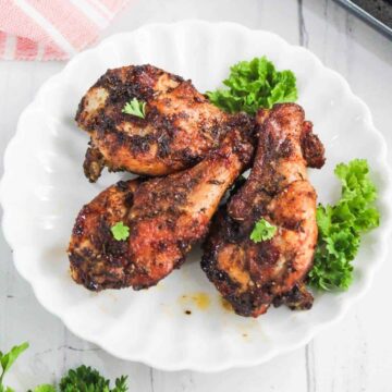 Three seasoned and cooked chicken drumsticks are arranged on a white plate, garnished with small sprigs of parsley and accompanied by fresh green lettuce.