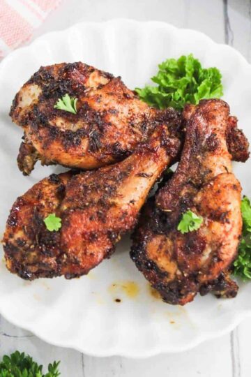 Three seasoned and cooked chicken drumsticks are arranged on a white plate, garnished with small sprigs of parsley and accompanied by fresh green lettuce.