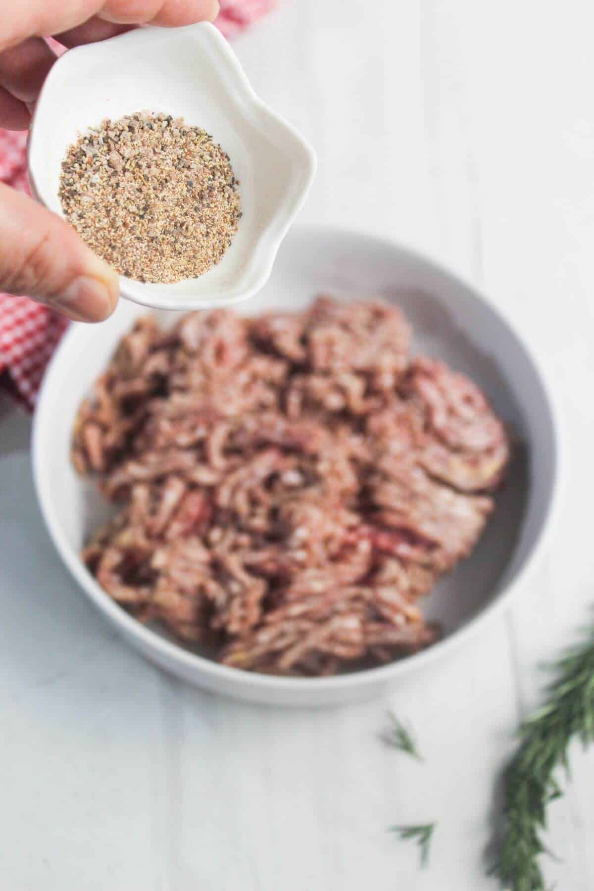 A hand holds a small bowl of seasoning above a larger bowl filled with raw ground meat on a white surface.
